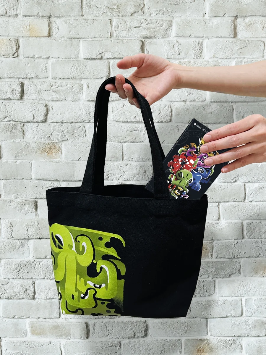 A hand carrying a black COC Canvas Bag with a green Cthulhu illustration and a game box inside